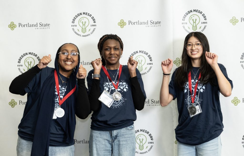 Three smiling students wearing medals and matching navy blue t-shirts pose with their fists raised in front of a Portland State University and Oregon MESA Day step-and-repeat backdrop for the MESA Day 2025 photo gallery.