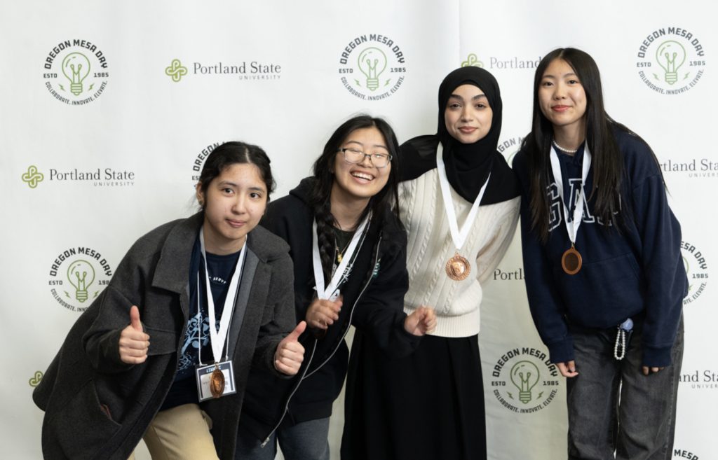 Four young women stand together smiling in front of a backdrop with Oregon MESA Day and Portland State University logos. Three wear medals, two give thumbs up, capturing a proud moment for the MESA Day 2025 photo gallery.