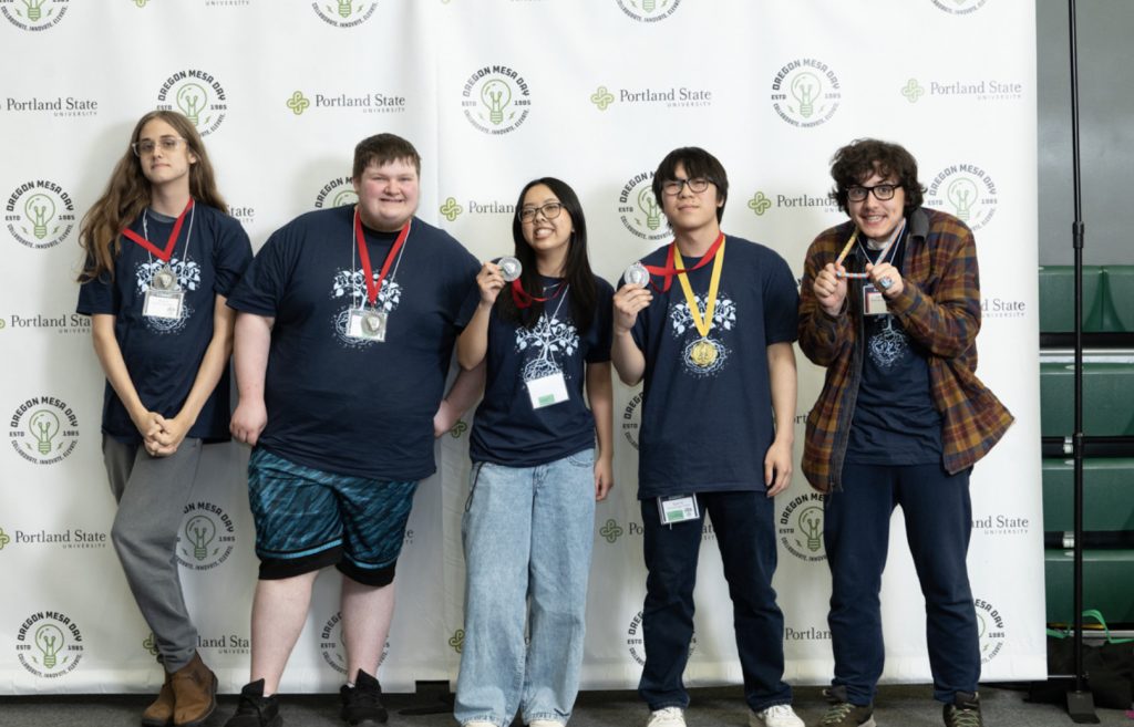 Five people stand in a row, wearing matching navy shirts and medals, smiling and making playful gestures at the mesa day 2025 photo gallery in front of a backdrop with Portland State and Oregon Media logos.