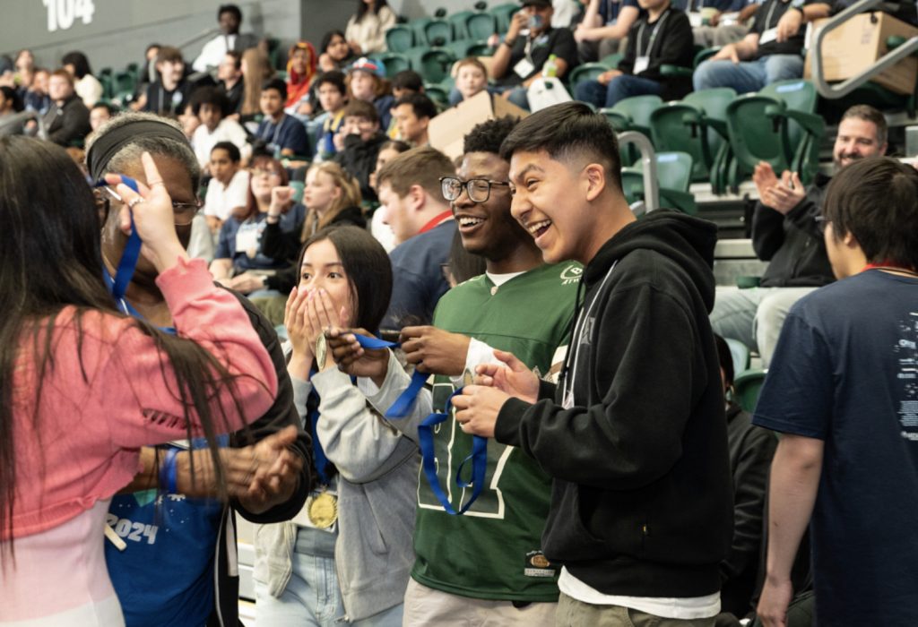 A group of excited young people stand in an auditorium, smiling and cheering as medals are handed out during Mesa Day 2025, while others in the background watch and applaud from green bleachers—a perfect moment for the photo gallery.