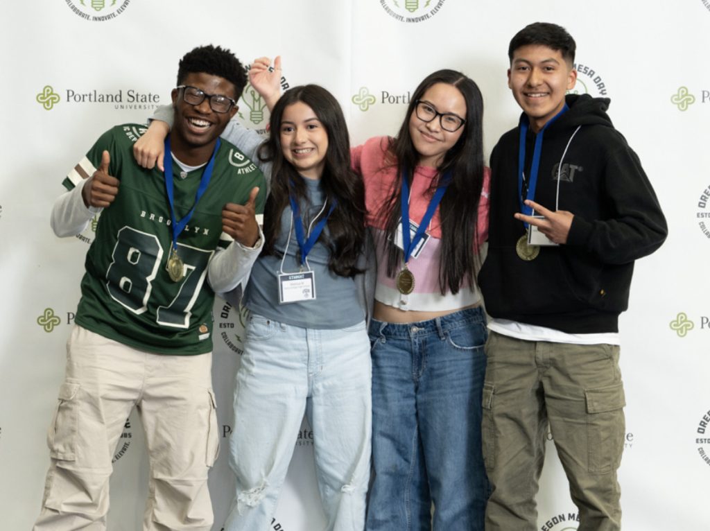 Four smiling students with medals around their necks stand arm-in-arm in front of a Portland State University backdrop, posing and making peace signs for the MESA Day 2025 photo gallery.