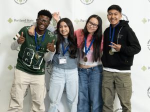 Four smiling students with medals around their necks stand arm-in-arm in front of a Portland State University backdrop, posing and making peace signs for the MESA Day 2025 photo gallery.