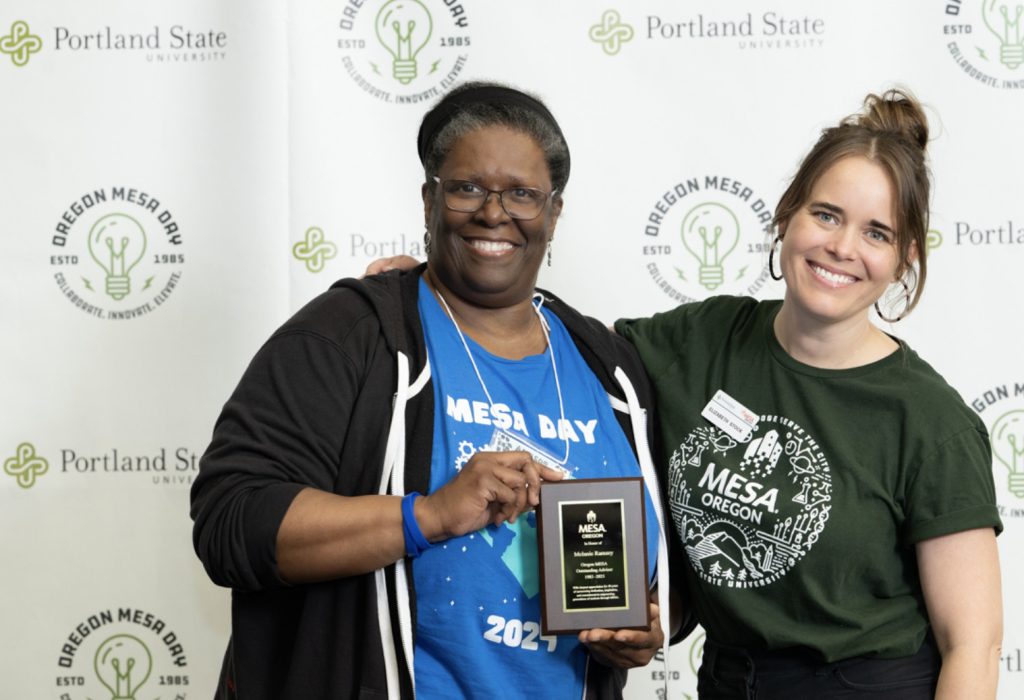 Two women smile at the camera; one holds a plaque that says MESA Day 2024. Both stand in front of a backdrop with Portland State University and Oregon MESA logos, perfect for the mesa day 2025 photo gallery.