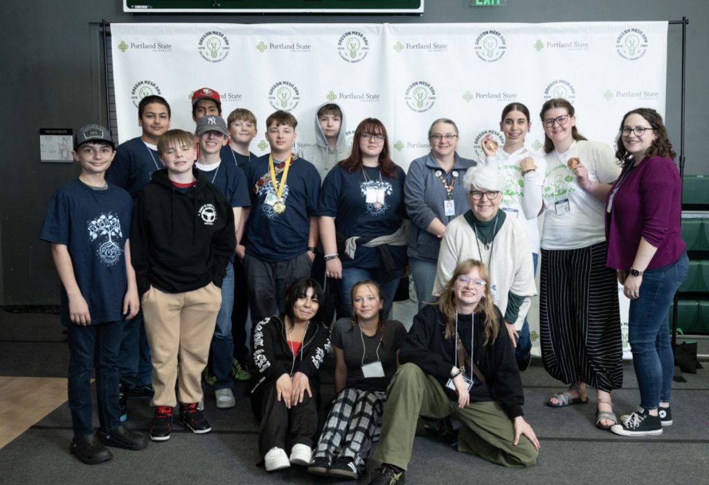A group of teenagers and a few adults pose together indoors in front of a white backdrop with logos for the Mesa Day 2025 photo gallery. Some are standing, some kneeling or sitting, and several are wearing medals and casual clothing.