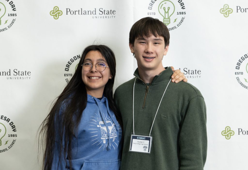 Two smiling young people pose together in front of a Portland State University and Oregon Day backdrop at the mesa day 2025 photo gallery. One wears glasses and a blue hoodie; the other sports a green sweater and a name tag.