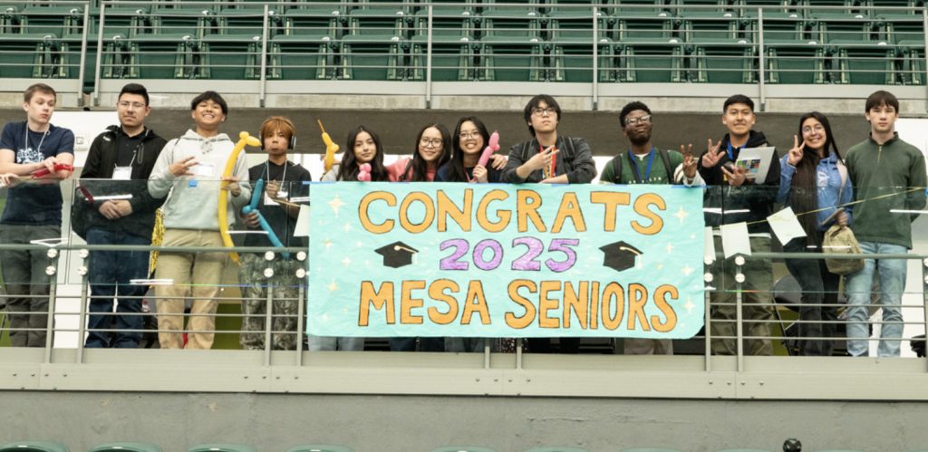 A group of students stand behind a balcony railing, holding a colorful sign that reads Congrats 2025 Mesa Seniors with graduation caps and stars drawn on it. They are smiling and posing for the camera in a stadium for the Mesa Day 2025 photo gallery.