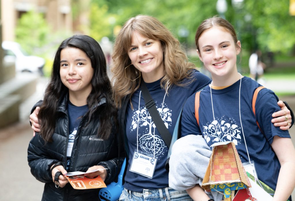 Three people wearing matching blue T-shirts with name tags smile outdoors at Mesa Day 2025. The person in the center has an arm around the others. One holds papers, another a colorful box. Trees and buildings are blurred in the photo gallery background.