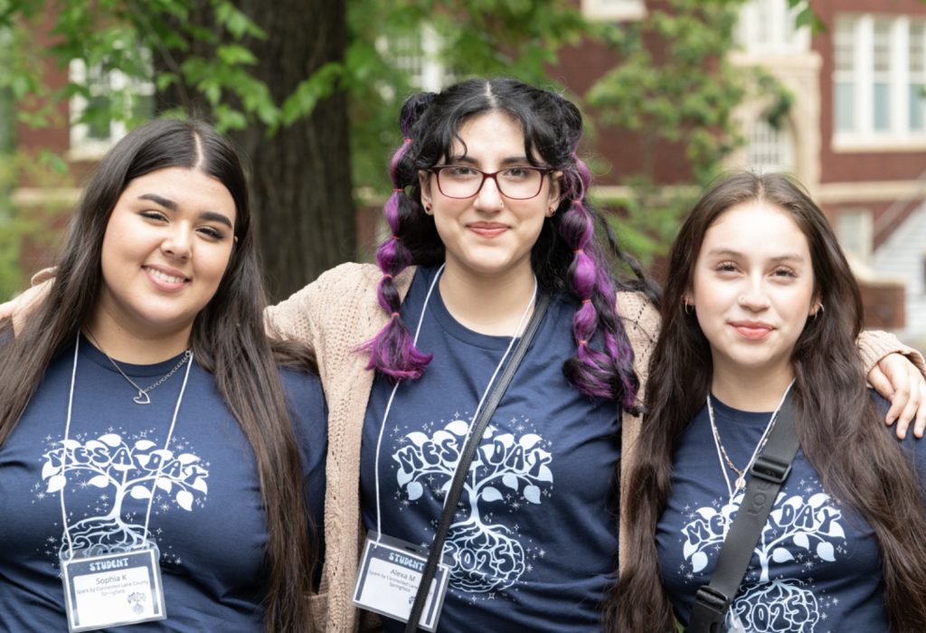 Three young women with long hair, wearing matching navy blue MESA Day 2023 shirts and name badges, stand closely together outdoors, smiling at the camera. This joyful moment is part of the MESA Day 2025 photo gallery.