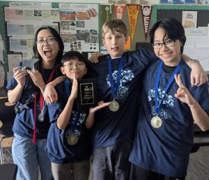 Meet the 2025 Oregon MESA State Champions: Four students in matching blue shirts and medals pose proudly in a classroom, holding trophies and celebrating. College pennants and educational posters decorate the walls behind them.
