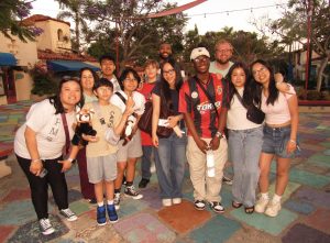 A group of thirteen people, both adults and children, stand close together smiling in an outdoor courtyard with colorful tiles and trees in the background. Some, proud Oregon MESA Students Shine at 2025 National Engineering Design Competition, are holding stuffed animals.