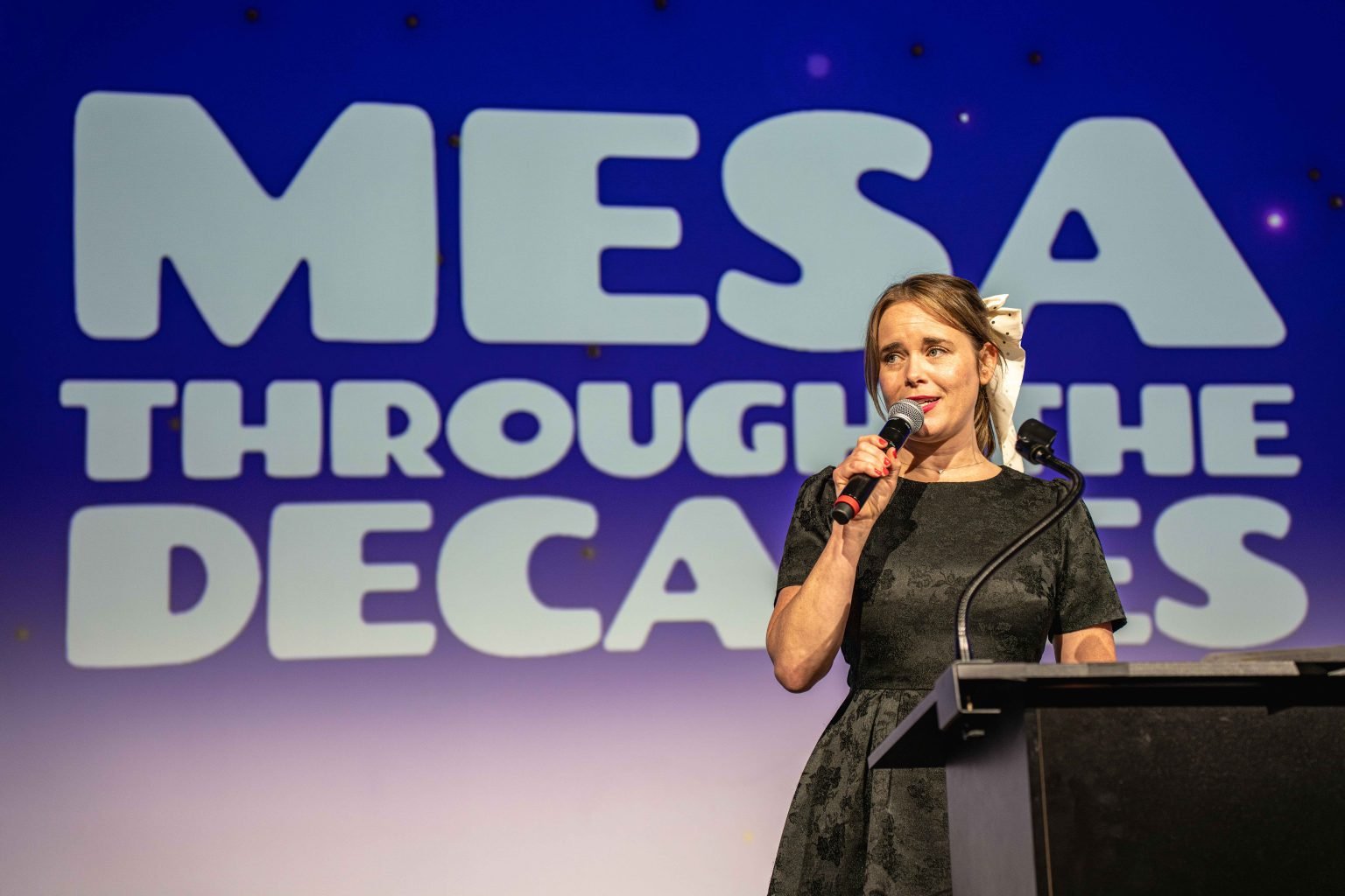 A woman in a black dress speaks into a microphone at a podium, standing in front of a screen displaying “MESA THROUGH THE DECADES” at the MESA Through the Decades 2025 Photo Gallery event.