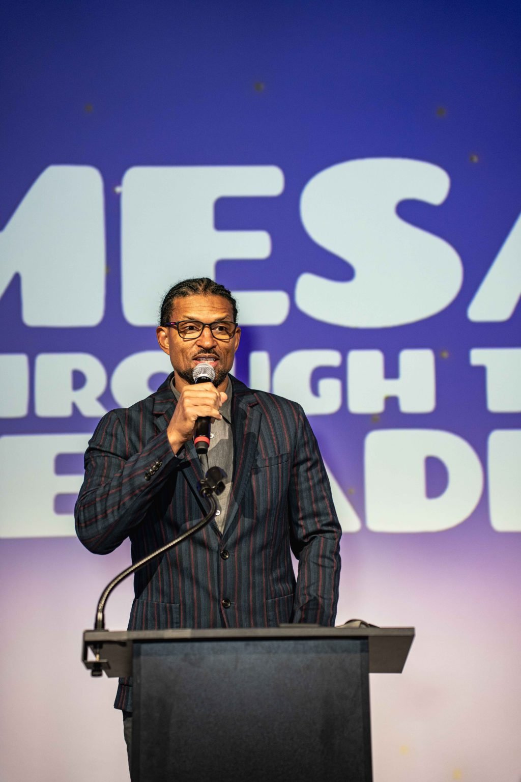 A man in a striped suit speaks into a microphone at a podium, standing in front of a blue and white backdrop with large, partially visible text at the MESA Through the Decades 2025 Photo Gallery.