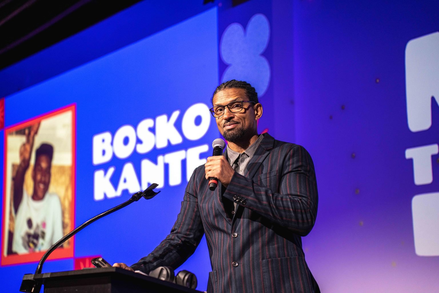 A man in a pinstripe suit and glasses speaks at a podium. Behind him, a large screen displays a childhood photo and the name Bosko Kante in bold white letters on a purple background for the MESA Through the Decades 2025 Photo Gallery event.
