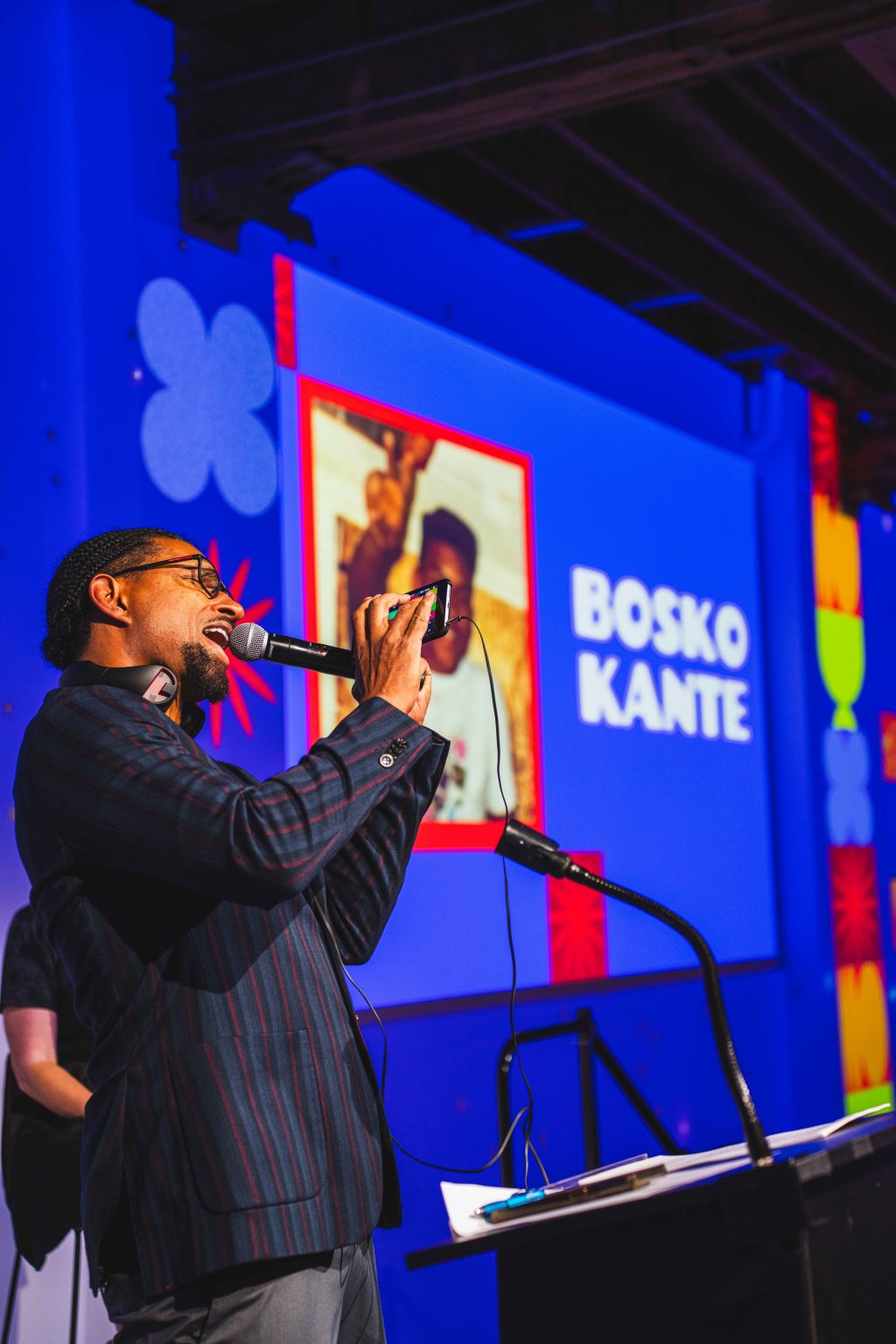 A man in glasses speaks and records with his phone on stage, as a large screen behind him displays Bosko Kante's name in bold white letters. The colorful background sets the scene for the MESA Through the Decades 2025 Photo Gallery event.