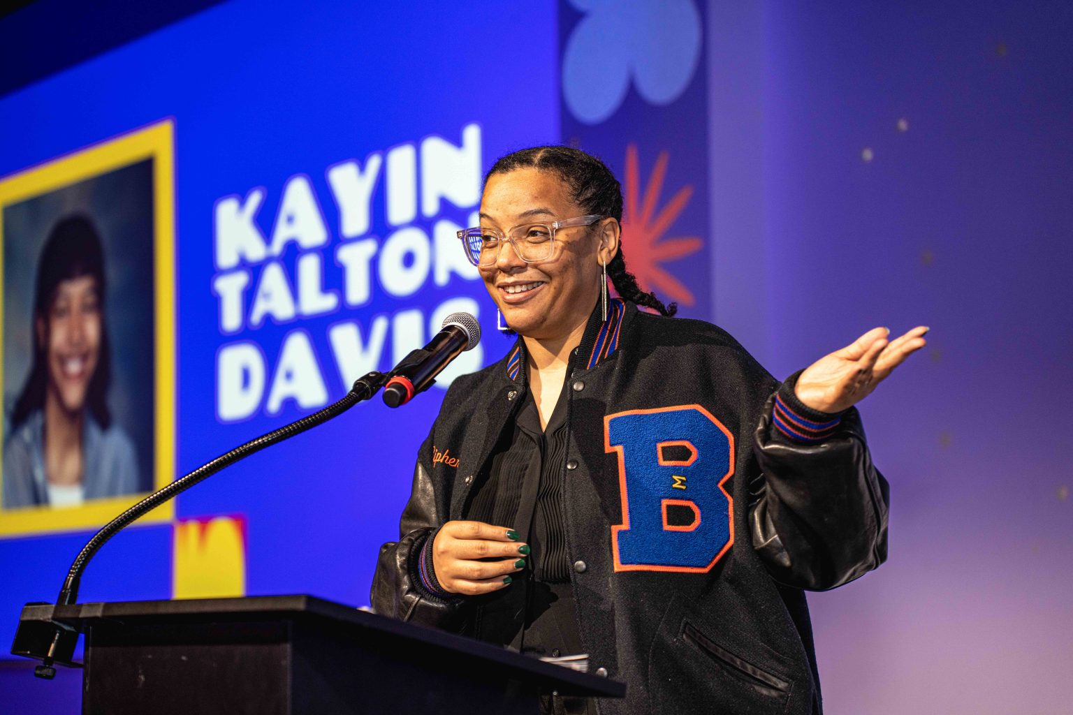A woman wearing glasses and a black letterman jacket speaks at a podium, smiling and gesturing. Behind her, a screen shows Kayin Talton Davis and a photo of a younger person at the MESA Through the Decades 2025 Photo Gallery.