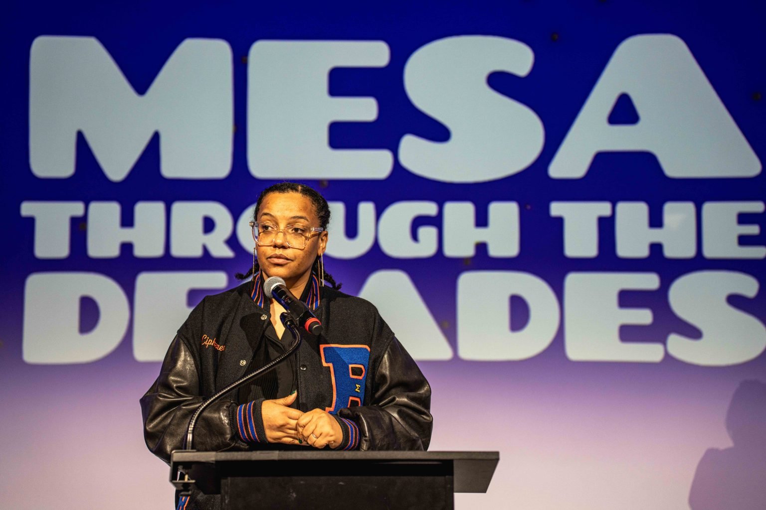 A person stands at a podium with a microphone, wearing glasses and a letterman jacket. Behind them, large text reads MESA THROUGH THE DECADES on a blue and white background—part of the MESA Through the Decades 2025 Photo Gallery.