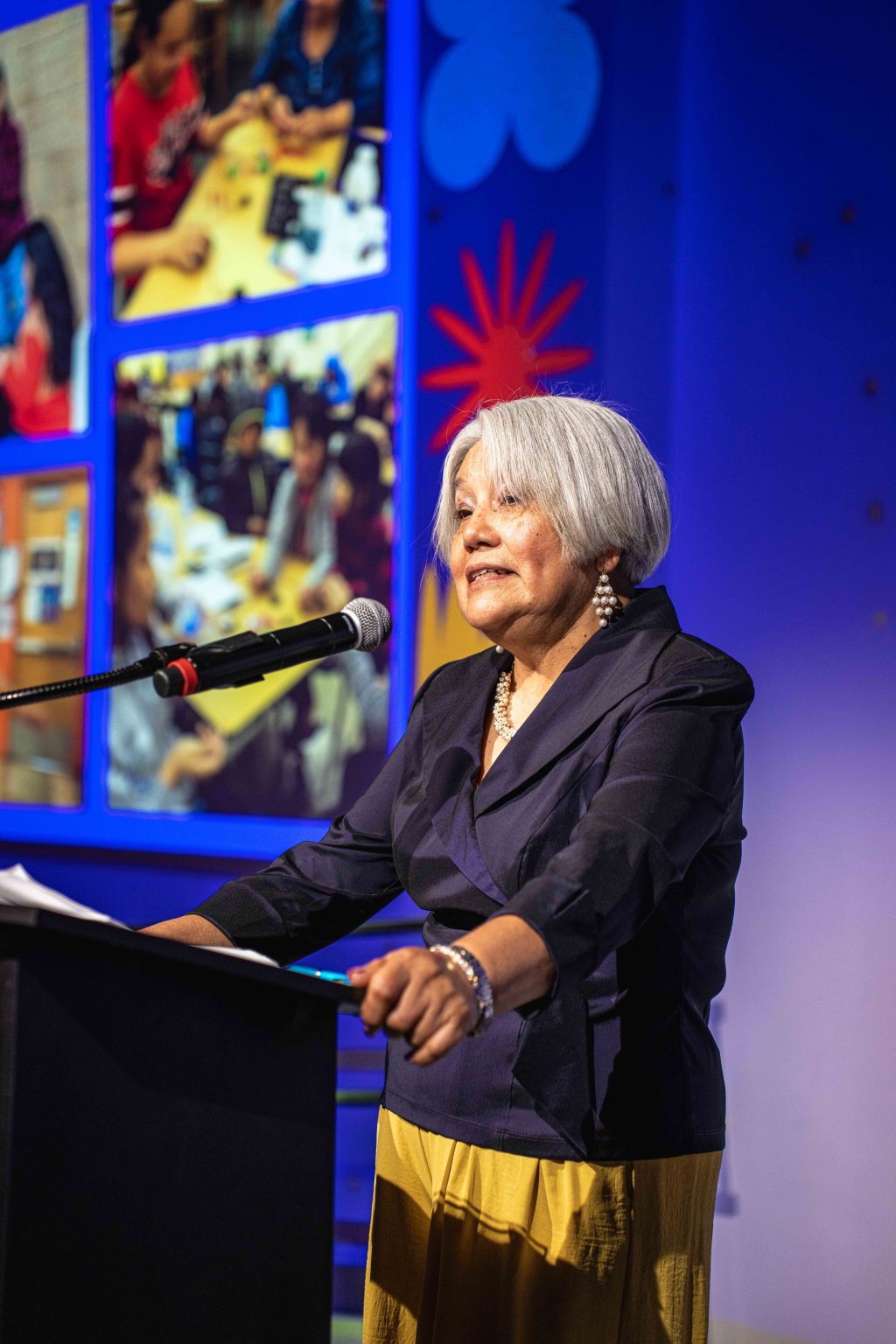 An older woman with short gray hair speaks at a podium with a microphone. She wears a dark jacket and yellow skirt. In the background, the MESA Through the Decades 2025 Photo Gallery features colorful photos on a blue wall.