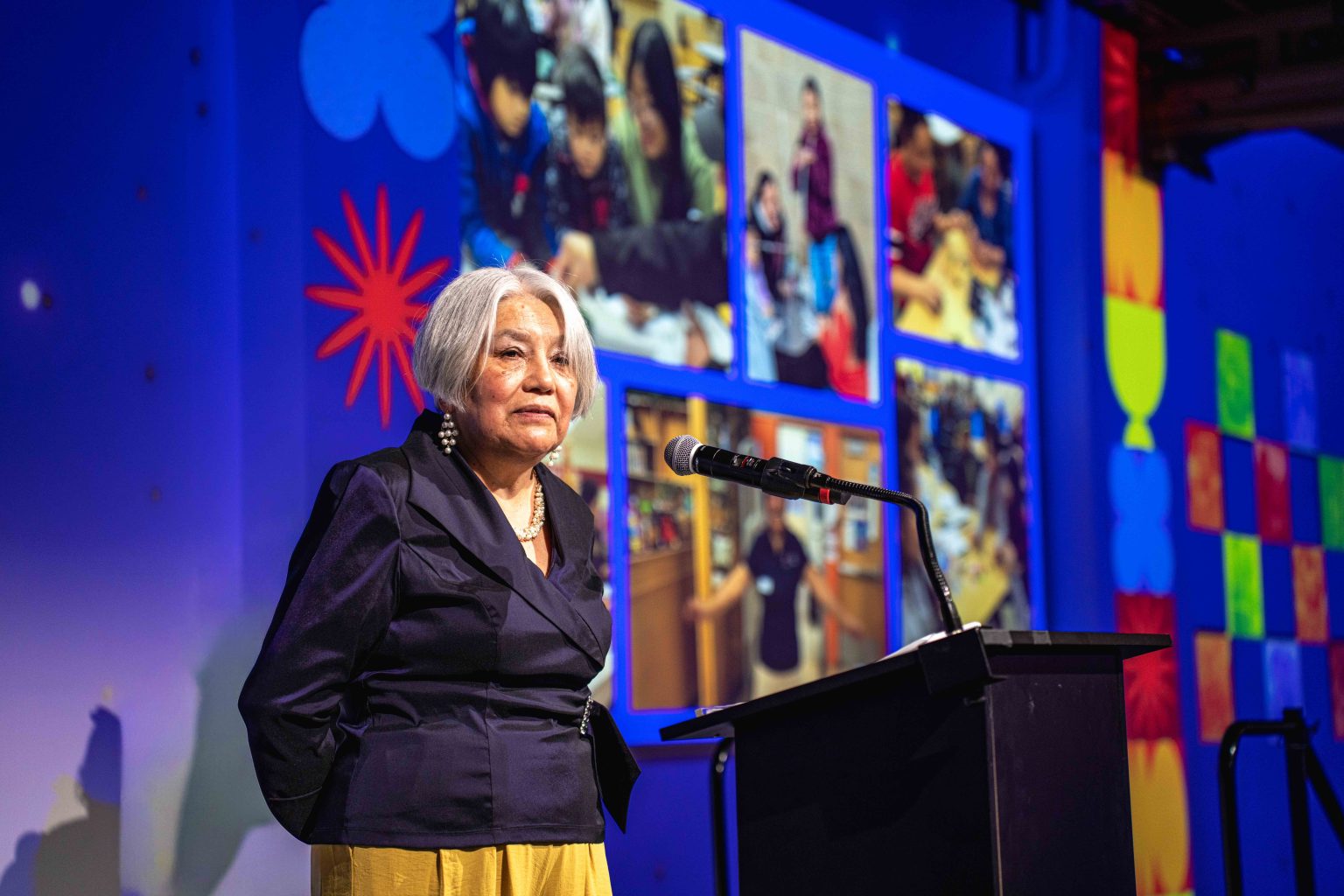 An older woman with gray hair stands at a podium speaking, with colorful walls and a screen behind her displaying the MESA Through the Decades 2025 Photo Gallery featuring people working together in group activities.