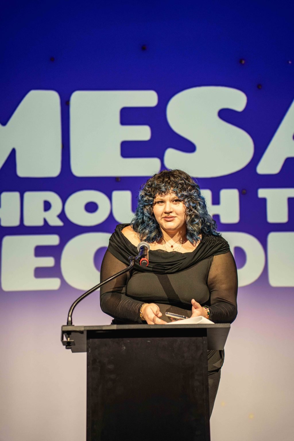 A person with curly blue hair stands at a podium, holding an award and speaking into a microphone, with large, partially visible text in the background and blue-purple lighting—part of the MESA Through the Decades 2025 Photo Gallery.