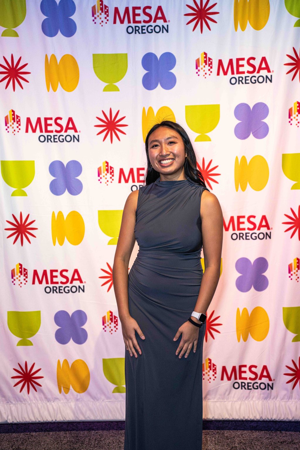 A woman in a sleeveless dark gray dress smiles while standing in front of a colorful backdrop with MESA OREGON logos and abstract shapes at the MESA Through the Decades 2025 Photo Gallery.