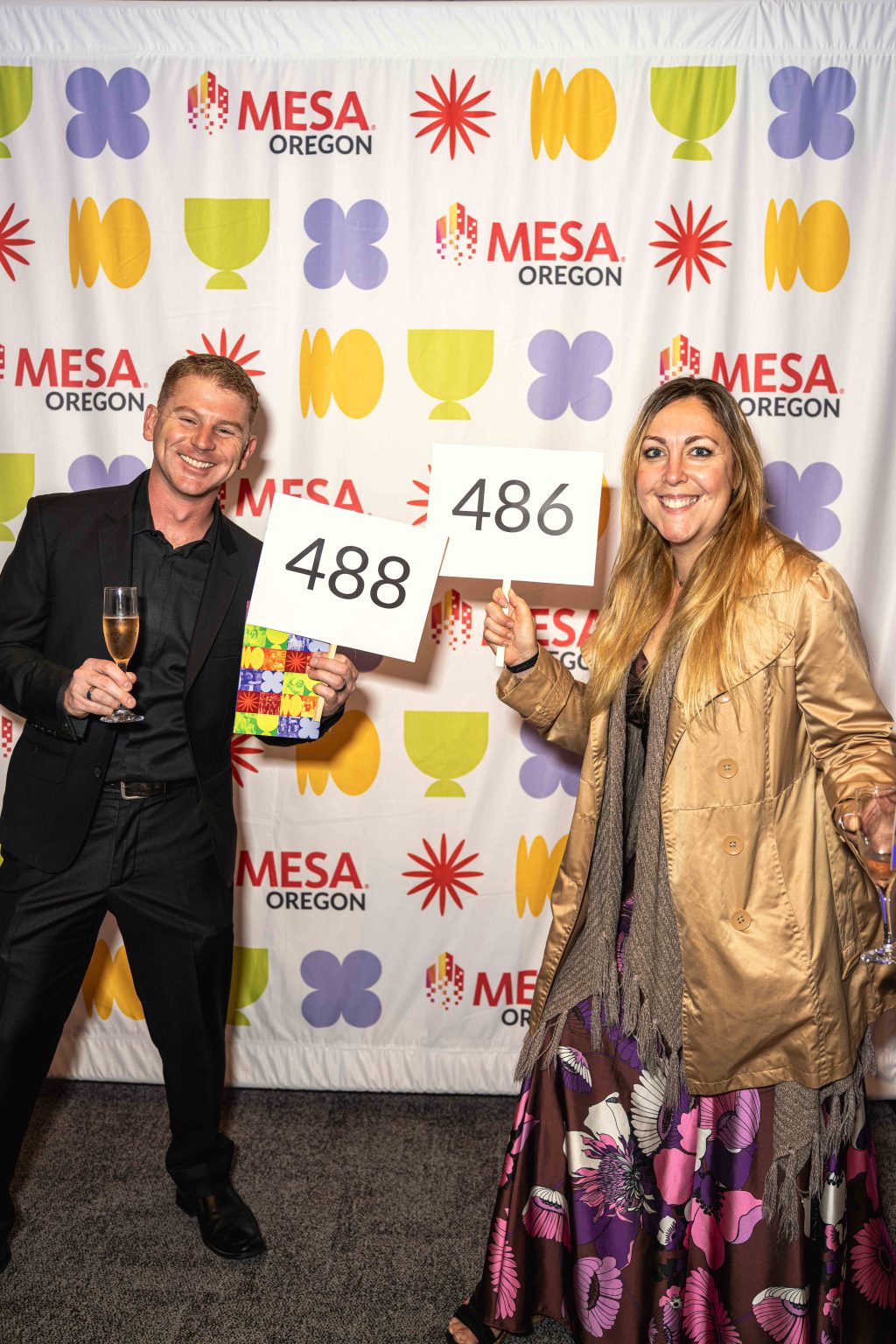A smiling man in a black suit holds a drink and a “488” card, while a smiling woman in a tan coat and floral dress holds a “486” card. They stand in front of the vibrant MESA Through the Decades 2025 Photo Gallery event backdrop.