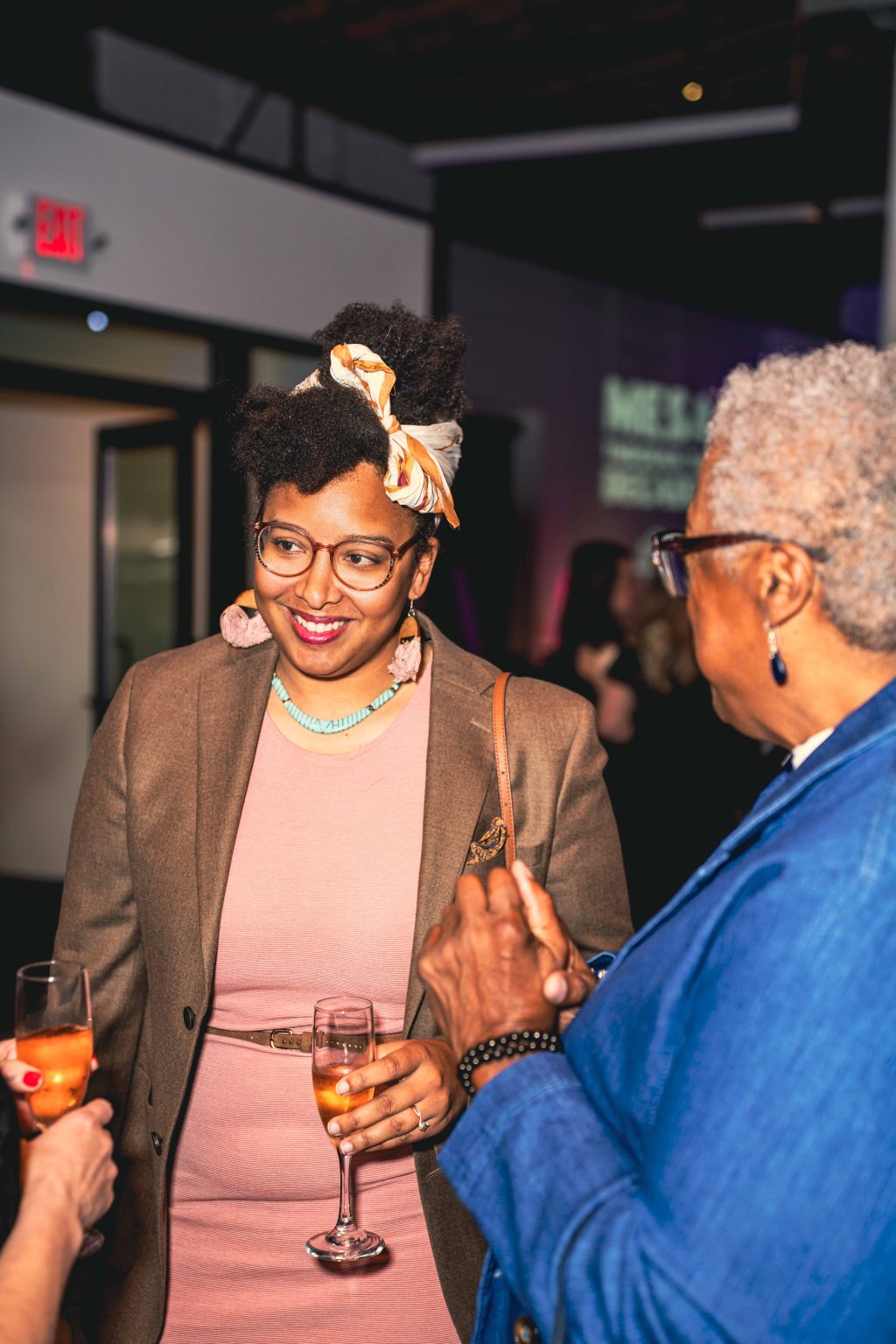 Two women stand indoors at a warmly lit social event, holding glasses of champagne and conversing. Captured for the MESA Through the Decades 2025 Photo Gallery, they wear chic outfits—a pink dress with a headscarf and a blue jacket.