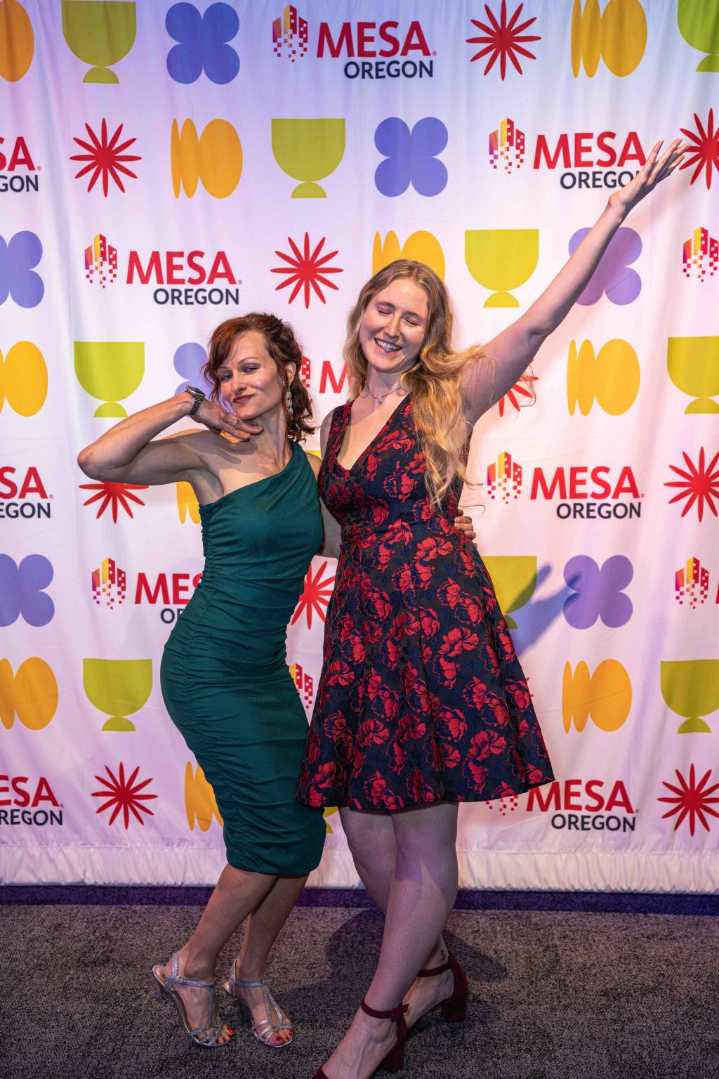 Two women pose playfully in front of a colorful MESA Oregon backdrop at the MESA Through the Decades 2025 Photo Gallery; both beaming, one wears a green dress and silver heels, the other a red floral dress and red shoes, arms raised in joy.