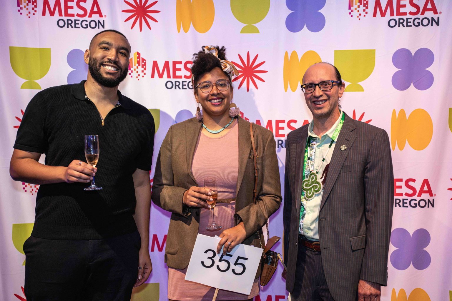 Three people smiling and holding glasses of champagne stand before a colorful MESA Oregon backdrop at the MESA Through the Decades 2025 Photo Gallery. The person in the center holds a paper with the number 355.
