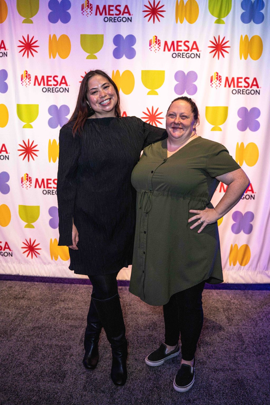 Two women smiling and posing together in front of a colorful backdrop with “MESA Oregon” logos and abstract shapes at the MESA Through the Decades 2025 Photo Gallery. One wears a black dress and boots; the other, a green dress and sneakers.
