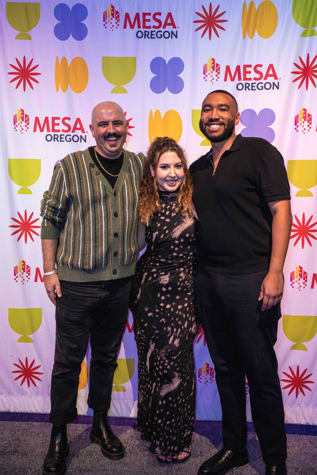 Three people stand smiling together in front of a colorful MESA Oregon backdrop at the MESA Through the Decades 2025 Photo Gallery. The person on the left wears a striped cardigan, the middle wears a black patterned dress, and the right wears black shirt and pants.