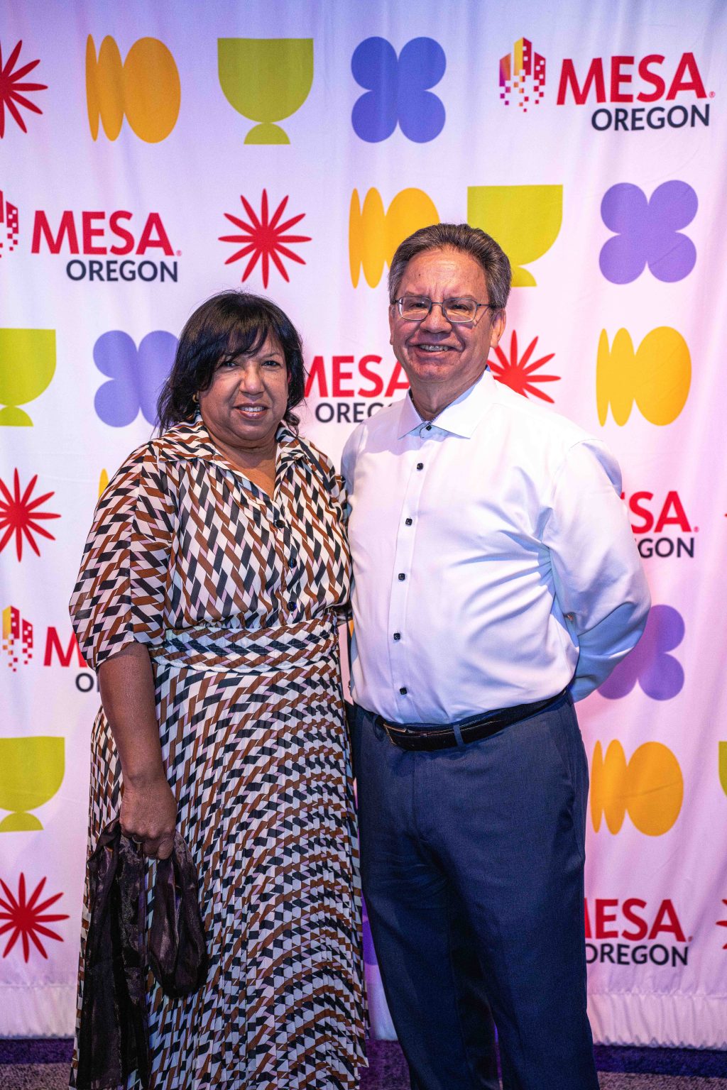 A woman in a patterned dress and a man in a white shirt pose together, smiling, in front of the colorful MESA Through the Decades 2025 Photo Gallery step-and-repeat banner with geometric shapes.