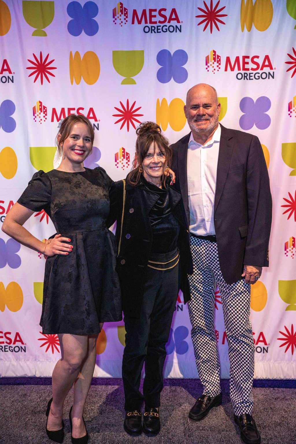 Three people stand smiling in front of a colorful MESA Through the Decades 2025 Photo Gallery backdrop. The two women wear black outfits, while the man sports a dark blazer, white shirt, and patterned pants.