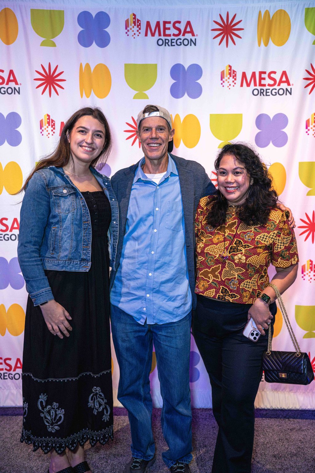 Three people smile and pose together in front of a colorful backdrop with MESA Oregon logos and geometric patterns, captured for the MESA Through the Decades 2025 Photo Gallery. The group includes two women and one man standing in the center.