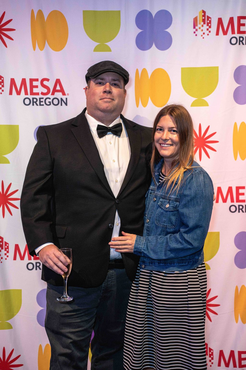 A man in a black suit and bow tie stands next to a woman in a denim jacket and striped dress, smiling, in front of the colorful MESA Through the Decades 2025 Photo Gallery step-and-repeat backdrop. The man holds a drink.