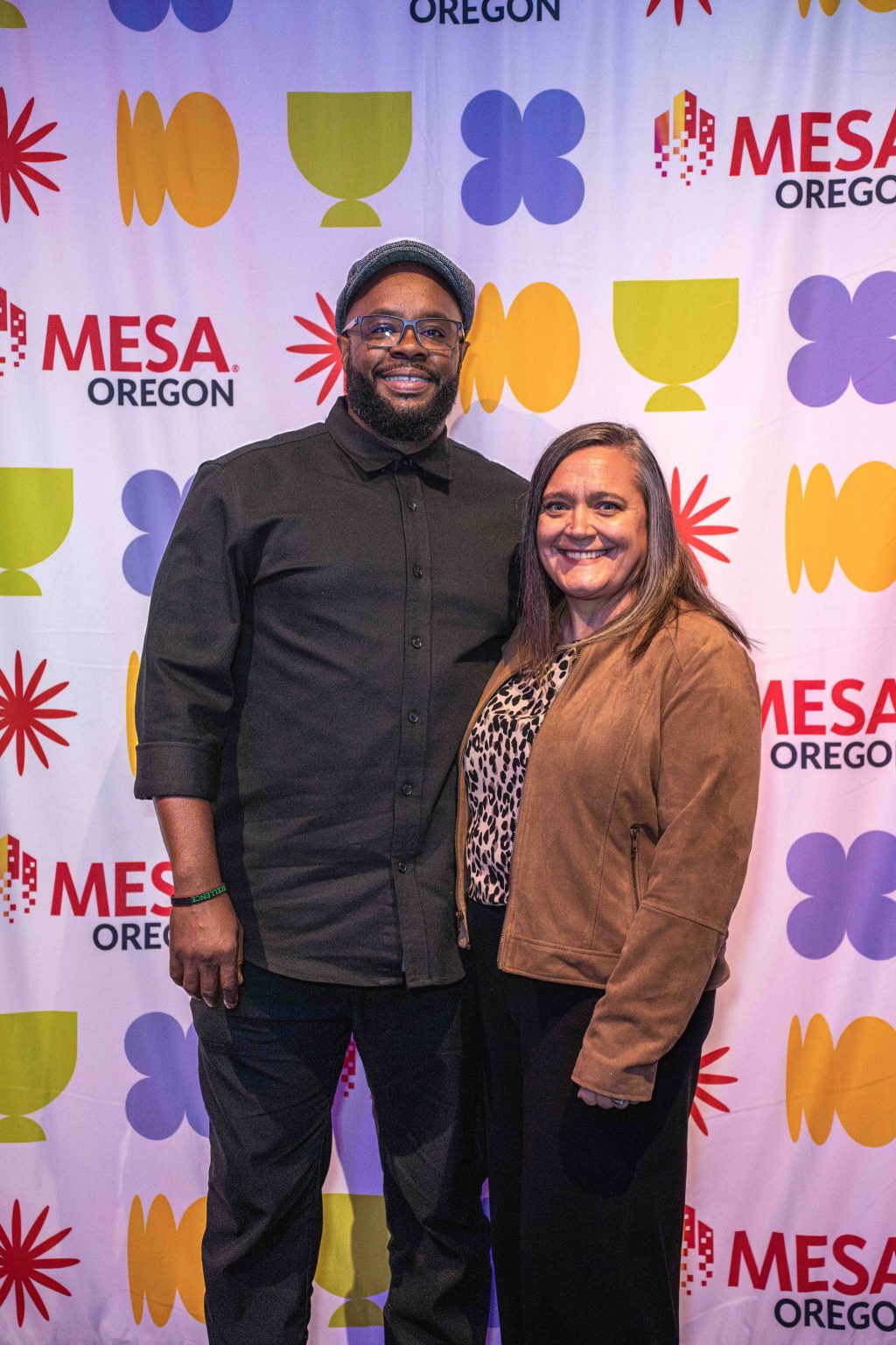 A man and woman stand smiling together in front of a colorful backdrop with the words MESA Oregon and various abstract shapes at the MESA Through the Decades 2025 Photo Gallery.