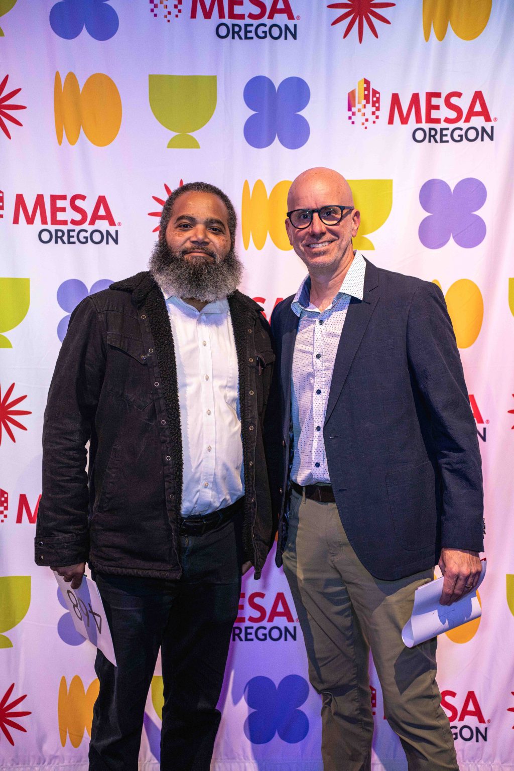 Two men stand side by side, smiling at the camera in front of a colorful MESA Oregon step-and-repeat backdrop. Both are holding papers and wearing jackets, capturing a moment for the MESA Through the Decades 2025 Photo Gallery.