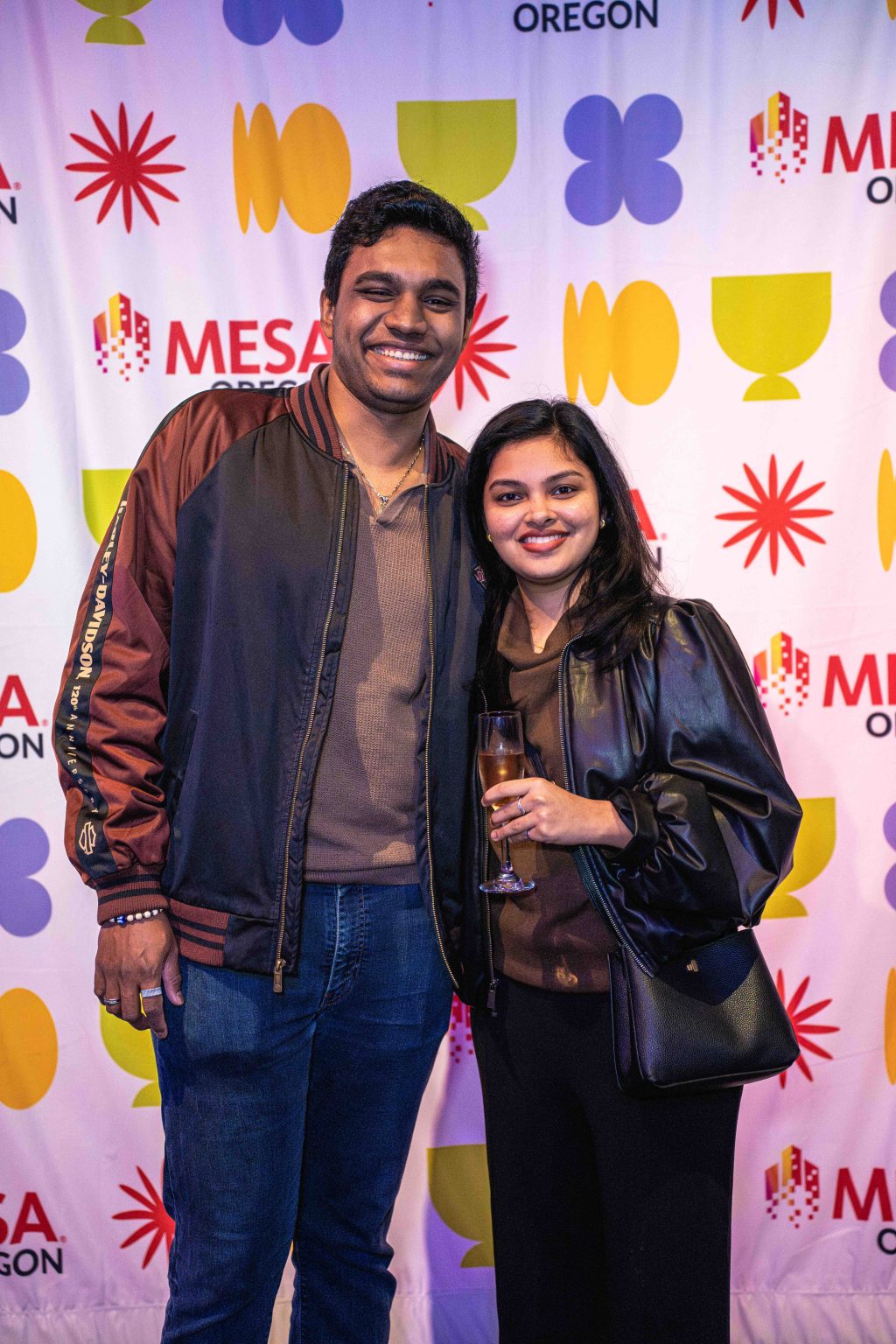 A smiling man and woman stand together in front of a colorful backdrop with geometric shapes and the words MESA Oregon. The woman holds a drink as they pose for the MESA Through the Decades 2025 Photo Gallery, both in casual jackets.