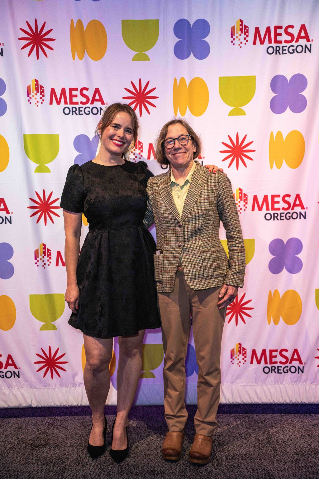 Two people stand smiling in front of a colorful backdrop with geometric shapes and the words MESA OREGON for the MESA Through the Decades 2025 Photo Gallery. One wears a black dress and heels; the other, glasses, a plaid blazer, light pants, and brown shoes.