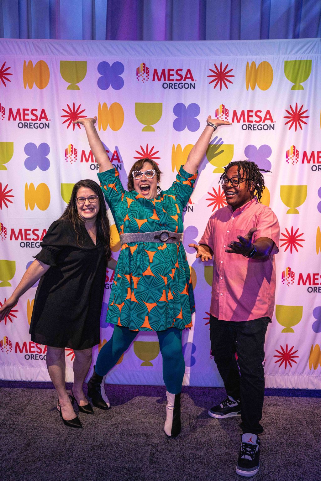 Three people pose enthusiastically in front of a colorful “MESA Oregon” step-and-repeat banner, creating a lively moment for the MESA Through the Decades 2025 Photo Gallery. The center person jumps with arms raised while the others smile and gesture energetically.