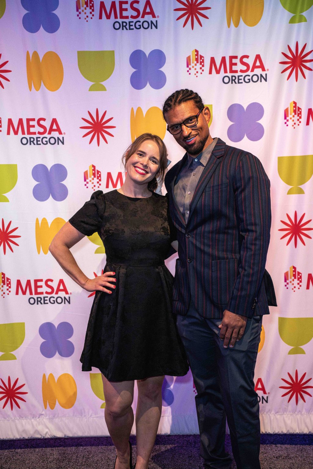 A woman in a black dress and a man in a striped suit jacket pose together, smiling, in front of the vibrant MESA Through the Decades 2025 Photo Gallery event backdrop.