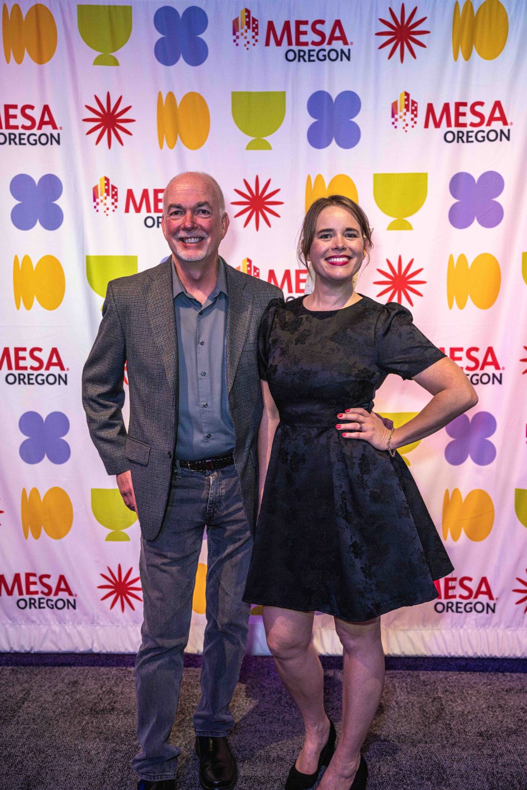 A man in a gray blazer and jeans stands next to a woman in a black dress, posing and smiling in front of a colorful “MESA Oregon” step-and-repeat backdrop at the MESA Through the Decades 2025 Photo Gallery.