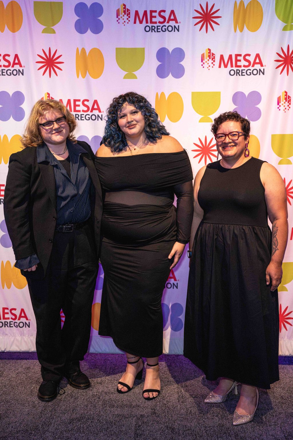 Three people stand smiling in front of a colorful step-and-repeat MESA Oregon backdrop at the MESA Through the Decades 2025 Photo Gallery. One wears a black suit, one a black off-the-shoulder dress, and the third a sleeveless black dress.