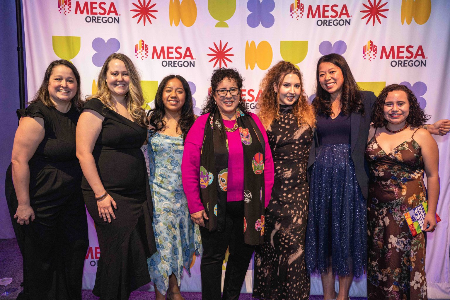 Seven women stand together smiling in front of a colorful step-and-repeat backdrop with “MESA Oregon” and abstract shapes, dressed in formal or semi-formal attire for the MESA Through the Decades 2025 Photo Gallery event.