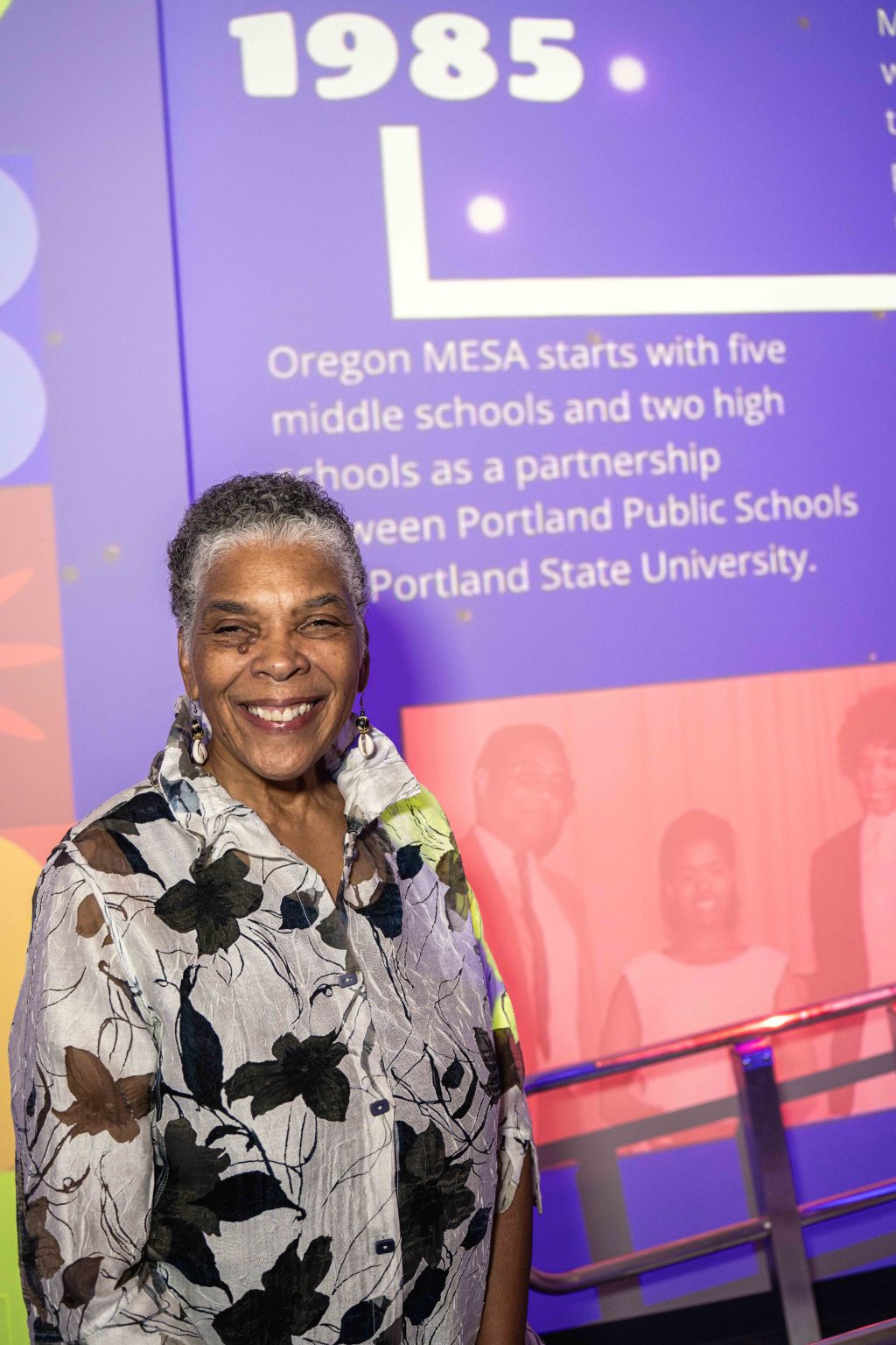 A smiling woman with short gray hair stands in front of a colorful background featuring the text “1985” and information about Oregon MESAs partnership with Portland Public Schools and Portland State University for the MESA Through the Decades 2025 Photo Gallery.