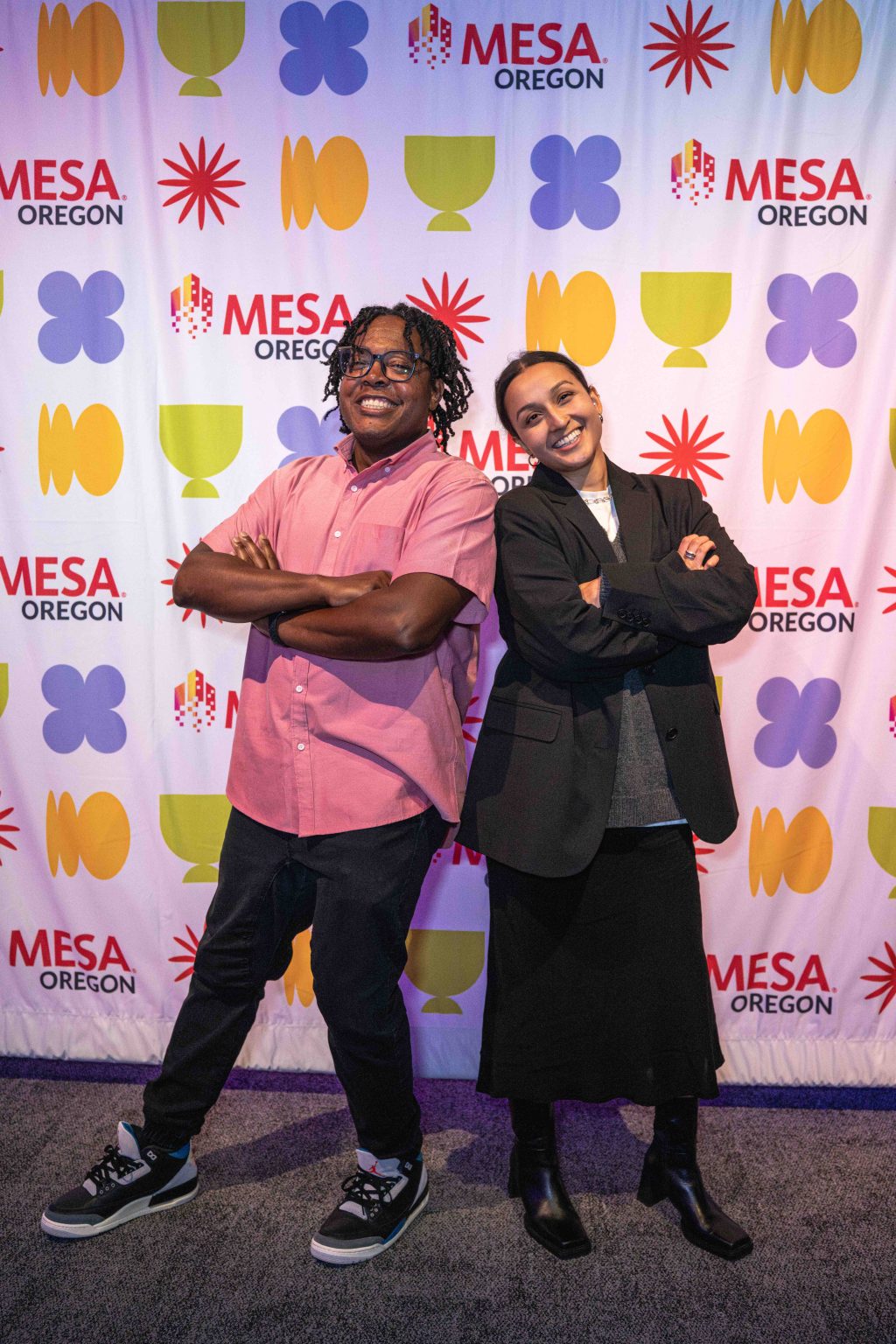 Two people stand back to back, smiling with arms crossed in front of a colorful Mesa Oregon step-and-repeat banner at the MESA Through the Decades 2025 Photo Gallery event. One wears a pink shirt and black pants, the other a black blazer and skirt.