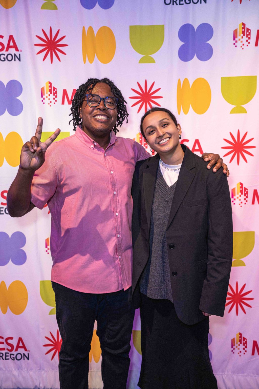 Two people pose and smile in front of a colorful backdrop with various logos at the MESA Through the Decades 2025 Photo Gallery. One wears a pink shirt and glasses, making a peace sign; the other sports a dark blazer and gray sweater.