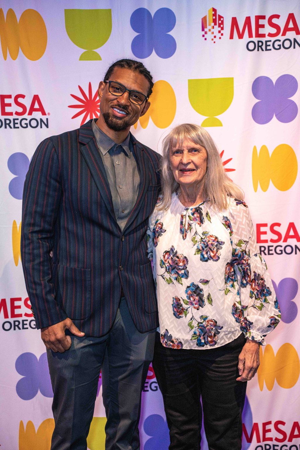 A man in a striped suit and glasses stands next to an older woman in a floral blouse. They are smiling in front of a colorful MESA Through the Decades 2025 Photo Gallery backdrop with Mesa Oregon and abstract shapes.