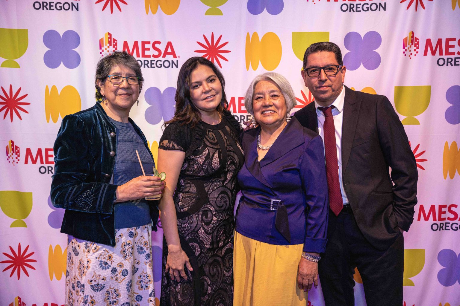 Four people stand together, dressed formally and smiling at the camera in front of a colorful backdrop featuring MESA OREGON, geometric shapes, and the MESA Through the Decades 2025 Photo Gallery.