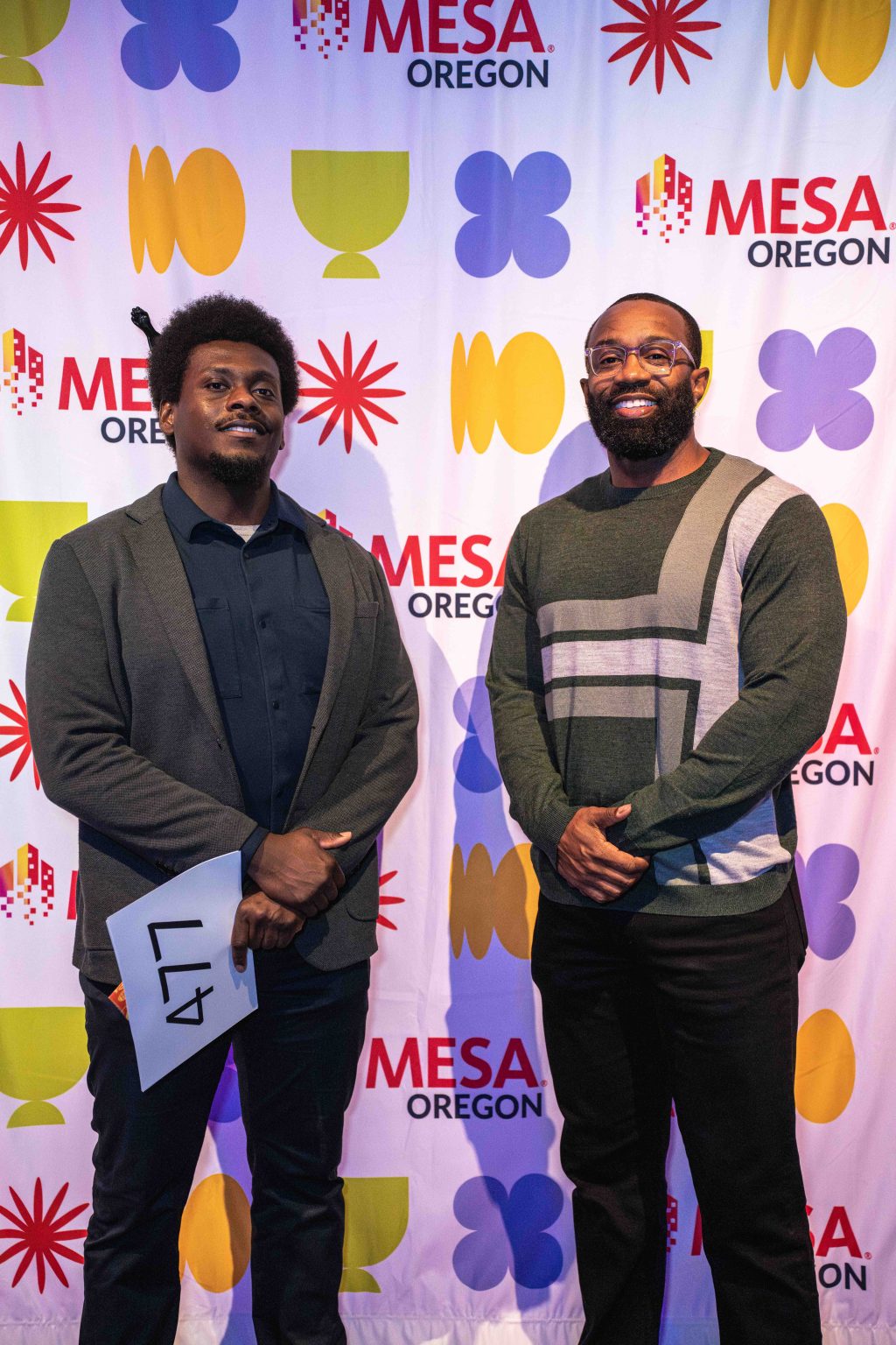 Two men stand and smile in front of a colorful MESA Oregon step-and-repeat banner. One holds a paper with the number 47. Both are dressed in dark pants and blazers, capturing a moment for the MESA Through the Decades 2025 Photo Gallery.
