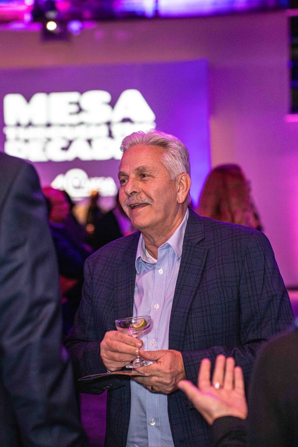 A man with gray hair and a mustache, dressed in a blue shirt and plaid blazer, holds a drink and smiles at the MESA Through the Decades 2025 Photo Gallery event. People and blurred “MESA” signage appear in the purple-lit background.